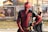 A person in a red and black superhero costume poses confidently with hands on hips in an outdoor setting. The background features blurred banners and a person wearing sunglasses standing casually.