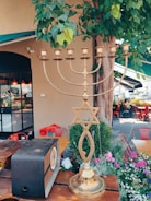 A brass menorah with a Star of David sits on a wooden table amidst a garden setting with lush greenery and colorful flowers. In the background, there is an old-fashioned radio, some red chairs, and a building with a black-framed window and awning.
