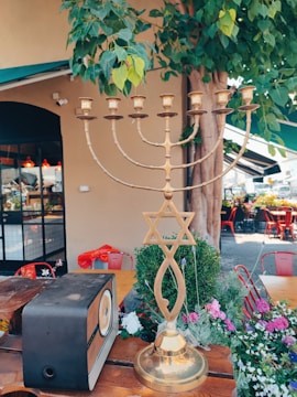 A brass menorah with a Star of David sits on a wooden table amidst a garden setting with lush greenery and colorful flowers. In the background, there is an old-fashioned radio, some red chairs, and a building with a black-framed window and awning.