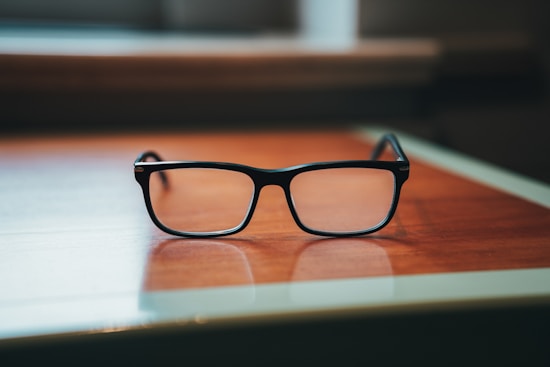 A pair of black-rimmed eyeglasses rests on a wooden table with a soft depth of field, creating a blurred background. The lighting is warm, highlighting the glasses and the texture of the table surface.