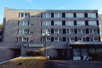 A multi-story building with a concrete facade featuring evenly spaced windows and a sign that reads 'Störtebeker' above a lower level entrance. A small garden or planted area is visible in the foreground, and light and shadow play on the building's surface adding depth to the structure. Two spherical outdoor lamps on tall poles stand near the entrance, with a sign to the right indicating a garden terrace.