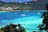 A picturesque view of a tranquil bay with turquoise waters, surrounded by lush green hills and dotted with several boats, including yachts. Luxury residences are visible along the shoreline in the background, and the scene is framed by tropical foliage in the foreground.