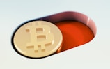 A gold coin embossed with the Bitcoin symbol is placed inside a red, pill-shaped recess. The background includes soft gradients of light colors, contrasting with the coin's golden hue.