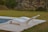 Two modern, curved white lounge chairs are positioned on a stone patio next to a swimming pool. The surrounding area is lush with green grass and mature trees, giving a sense of tranquility and relaxation.