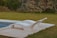 Two modern, curved white lounge chairs are positioned on a stone patio next to a swimming pool. The surrounding area is lush with green grass and mature trees, giving a sense of tranquility and relaxation.