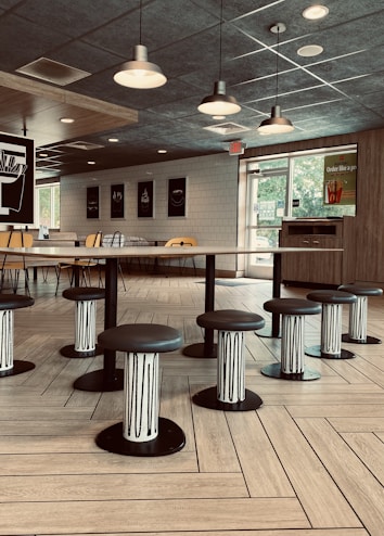 A modern cafe interior with a series of round stools featuring black and white patterned bases. The room has wooden flooring with a chevron pattern and is lit by a series of pendant lights hanging from the ceiling. The walls are adorned with minimalist art prints and there is a large window letting in natural light. The overall ambiance is clean and contemporary.
