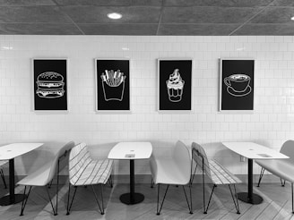 A modern café setting featuring white walls covered with white tiles and minimalist black-and-white artwork of food items such as a burger, fries, ice cream, and a cup of coffee. There are sleek white tables and a combination of white chairs and metal wire benches arranged neatly in front of the artwork. The overall theme is minimalistic and monochromatic.