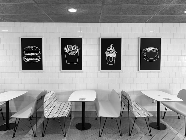 A modern café setting featuring white walls covered with white tiles and minimalist black-and-white artwork of food items such as a burger, fries, ice cream, and a cup of coffee. There are sleek white tables and a combination of white chairs and metal wire benches arranged neatly in front of the artwork. The overall theme is minimalistic and monochromatic.