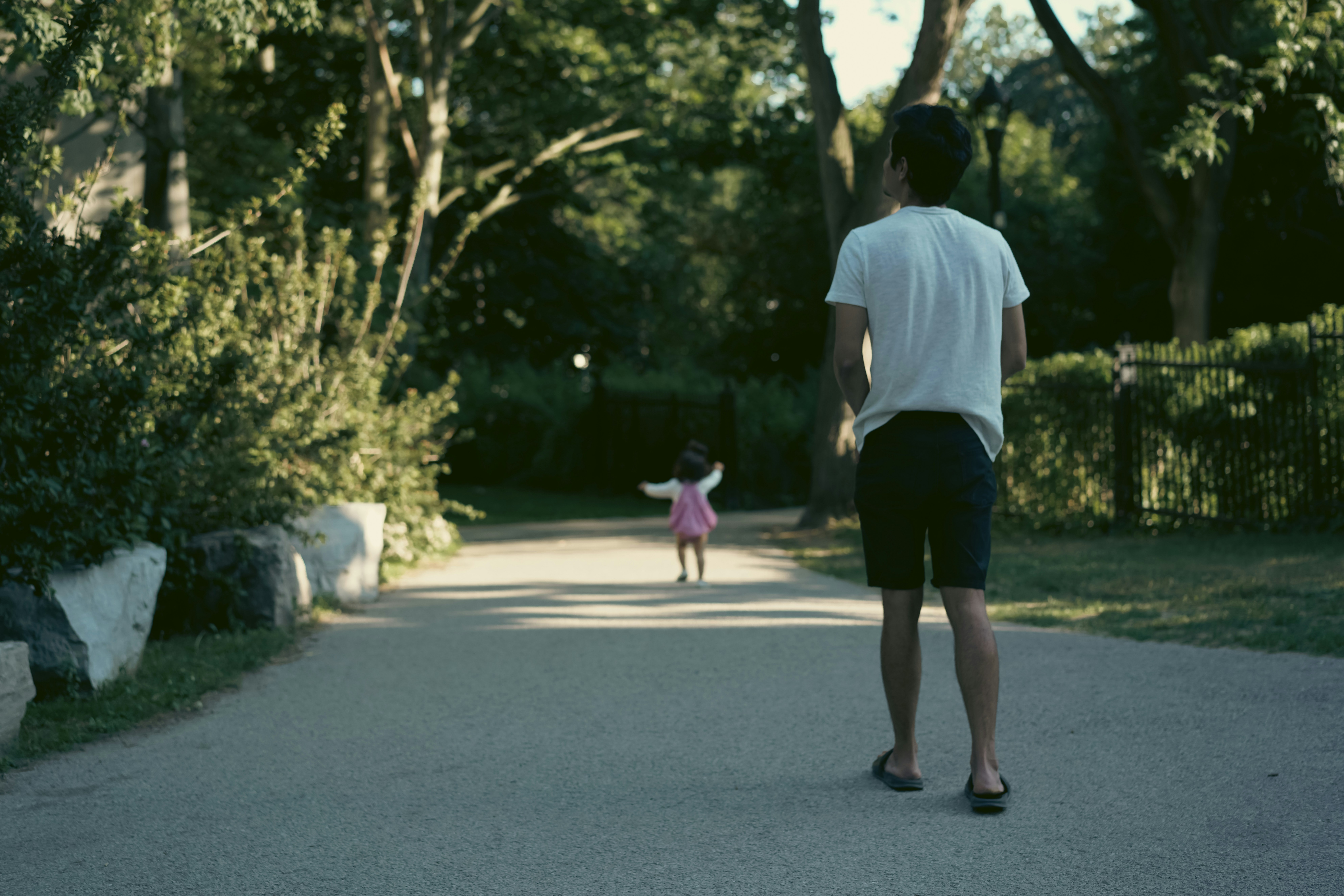 A small girl in a pink dress and white jacket runs joyfully down a wide paved path lined with greenery, her arms in the air, while her father, in a white t-shirt and shorts, walks behind her, looking upward