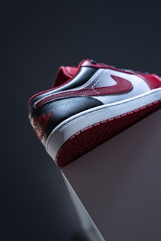 A close-up of a sneaker with a red, white, and gray color scheme. The angle highlights the side panel, heel, and sole details, showcasing the texture and design elements.