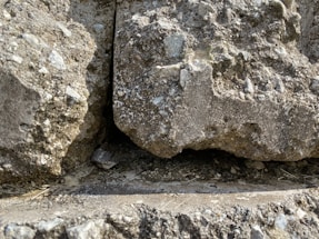 The image displays a close-up view of a rugged rock surface with a visible gap between two large stones. The rocks have a rough texture with small pebbles and uneven surfaces, and there is a shadowed area within the gap.