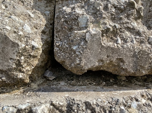 The image displays a close-up view of a rugged rock surface with a visible gap between two large stones. The rocks have a rough texture with small pebbles and uneven surfaces, and there is a shadowed area within the gap.