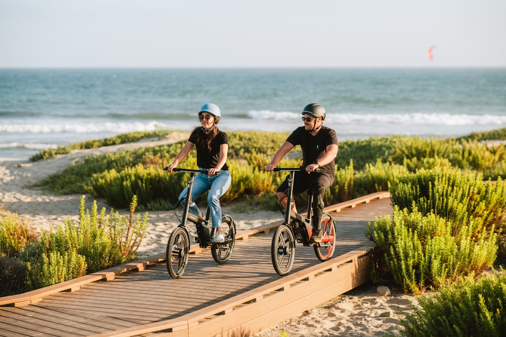 a couple of people riding bikes on a boardwalk by the beach