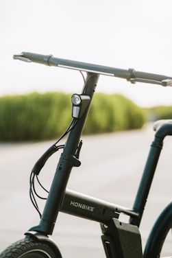 A close-up view of a sleek, modern bicycle featuring a straight handlebar with hand grips and a small front light mounted on the front frame tube. The bicycle has a minimalist design with visible wiring and a rectangular battery pack integrated into the frame. The background shows a softly blurred, natural landscape with green foliage.