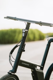 A close-up view of a sleek, modern bicycle featuring a straight handlebar with hand grips and a small front light mounted on the front frame tube. The bicycle has a minimalist design with visible wiring and a rectangular battery pack integrated into the frame. The background shows a softly blurred, natural landscape with green foliage.