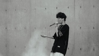 A person with curly hair is performing with a microphone against a textured concrete wall. The individual is wearing glasses and a dark oversized shirt with visible white lettering on the sleeve.