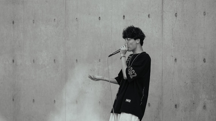 A person with curly hair is performing with a microphone against a textured concrete wall. The individual is wearing glasses and a dark oversized shirt with visible white lettering on the sleeve.