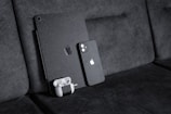 A black textured iPad with a matching iPhone resting against a dark fabric sofa. A pair of white wireless earbuds in a case is attached to the iPad. The devices have a honeycomb pattern skin and feature the Apple logo.