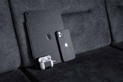 A black textured iPad with a matching iPhone resting against a dark fabric sofa. A pair of white wireless earbuds in a case is attached to the iPad. The devices have a honeycomb pattern skin and feature the Apple logo.