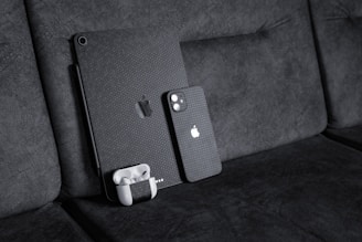 A black textured iPad with a matching iPhone resting against a dark fabric sofa. A pair of white wireless earbuds in a case is attached to the iPad. The devices have a honeycomb pattern skin and feature the Apple logo.