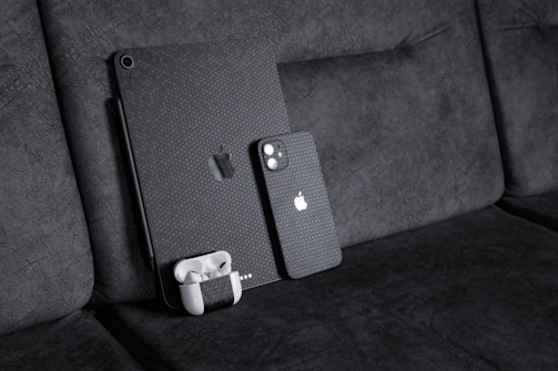 A black textured iPad with a matching iPhone resting against a dark fabric sofa. A pair of white wireless earbuds in a case is attached to the iPad. The devices have a honeycomb pattern skin and feature the Apple logo.