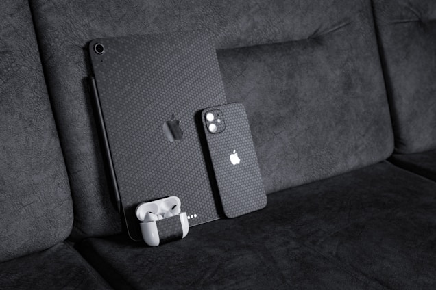 A black textured iPad with a matching iPhone resting against a dark fabric sofa. A pair of white wireless earbuds in a case is attached to the iPad. The devices have a honeycomb pattern skin and feature the Apple logo.