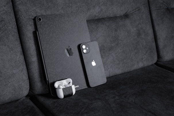 A black textured iPad with a matching iPhone resting against a dark fabric sofa. A pair of white wireless earbuds in a case is attached to the iPad. The devices have a honeycomb pattern skin and feature the Apple logo.