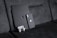 A black textured iPad with a matching iPhone resting against a dark fabric sofa. A pair of white wireless earbuds in a case is attached to the iPad. The devices have a honeycomb pattern skin and feature the Apple logo.