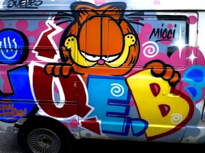 A colorful graffiti artwork on the side of a vehicle featuring a cartoon character with orange fur and a mischievous expression, peering over large, stylized letters 'JUEB' painted in bold shades of blue, yellow, and red. The background is adorned with vibrant swirls, dots, and striking accents in pink and white.