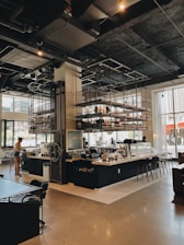 A modern cafe with industrial-style decor is shown, featuring high ceilings and exposed ductwork. The space includes a large counter area with seating, shelving with various items, and a barista preparing drinks. Natural light streams in through large windows, creating a warm ambiance. The room is spacious with contemporary furniture.