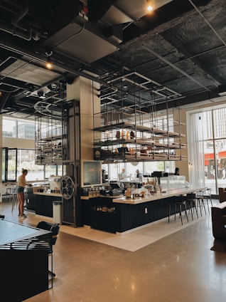 A modern cafe with industrial-style decor is shown, featuring high ceilings and exposed ductwork. The space includes a large counter area with seating, shelving with various items, and a barista preparing drinks. Natural light streams in through large windows, creating a warm ambiance. The room is spacious with contemporary furniture.
