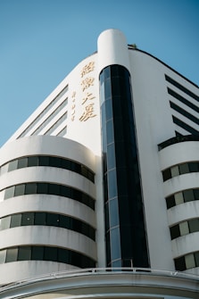 A modern, multi-story building featuring a white facade with large, dark-tinted windows. The design includes a prominent vertical architectural element, possibly housing an elevator or staircase. Gold-colored inscriptions are visible on the building's exterior.