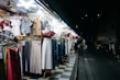 A street market stall displays a variety of clothing items, including dresses, blouses, pants, and jackets, all arranged on hangers and mannequins. The scene is illuminated by bright overhead lights, contrasting with the dark, empty street in the background where a few pedestrians are visible.