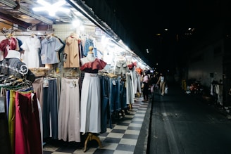 A street market stall displays a variety of clothing items, including dresses, blouses, pants, and jackets, all arranged on hangers and mannequins. The scene is illuminated by bright overhead lights, contrasting with the dark, empty street in the background where a few pedestrians are visible.