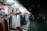 A street market stall displays a variety of clothing items, including dresses, blouses, pants, and jackets, all arranged on hangers and mannequins. The scene is illuminated by bright overhead lights, contrasting with the dark, empty street in the background where a few pedestrians are visible.