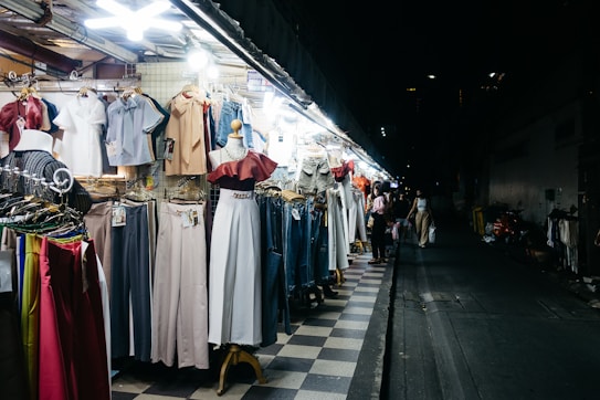 A street market stall displays a variety of clothing items, including dresses, blouses, pants, and jackets, all arranged on hangers and mannequins. The scene is illuminated by bright overhead lights, contrasting with the dark, empty street in the background where a few pedestrians are visible.