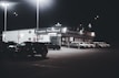 A dimly lit car dealership at night features a row of premium pre-owned cars parked in front of a building with bright lights. The building is adorned with various signs and has a 'Welcome' message visible. Multiple street lamps illuminate the parking lot.