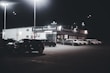 A dimly lit car dealership at night features a row of premium pre-owned cars parked in front of a building with bright lights. The building is adorned with various signs and has a 'Welcome' message visible. Multiple street lamps illuminate the parking lot.