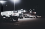 A dimly lit car dealership at night features a row of premium pre-owned cars parked in front of a building with bright lights. The building is adorned with various signs and has a 'Welcome' message visible. Multiple street lamps illuminate the parking lot.