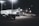 A dimly lit car dealership at night features a row of premium pre-owned cars parked in front of a building with bright lights. The building is adorned with various signs and has a 'Welcome' message visible. Multiple street lamps illuminate the parking lot.