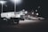 A dimly lit car dealership at night features a row of premium pre-owned cars parked in front of a building with bright lights. The building is adorned with various signs and has a 'Welcome' message visible. Multiple street lamps illuminate the parking lot.