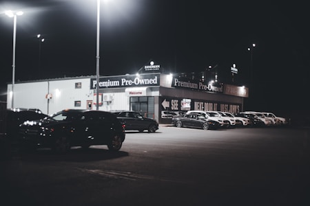A dimly lit car dealership at night features a row of premium pre-owned cars parked in front of a building with bright lights. The building is adorned with various signs and has a 'Welcome' message visible. Multiple street lamps illuminate the parking lot.