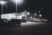 A dimly lit car dealership at night features a row of premium pre-owned cars parked in front of a building with bright lights. The building is adorned with various signs and has a 'Welcome' message visible. Multiple street lamps illuminate the parking lot.