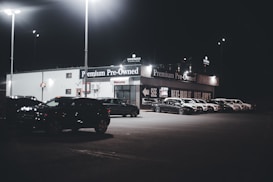 A dimly lit car dealership at night features a row of premium pre-owned cars parked in front of a building with bright lights. The building is adorned with various signs and has a 'Welcome' message visible. Multiple street lamps illuminate the parking lot.