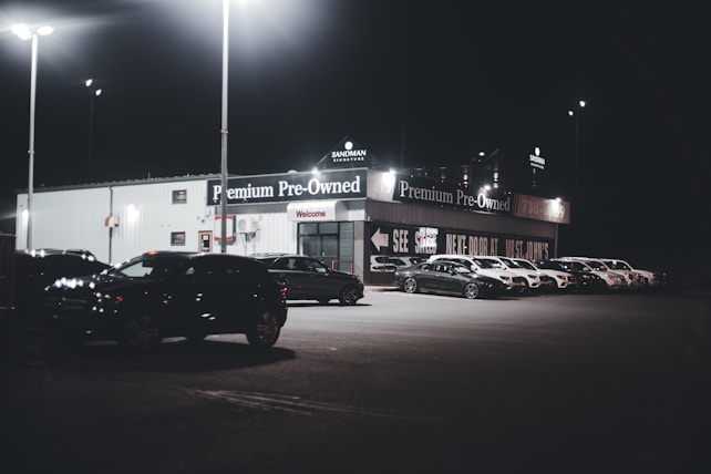 A dimly lit car dealership at night features a row of premium pre-owned cars parked in front of a building with bright lights. The building is adorned with various signs and has a 'Welcome' message visible. Multiple street lamps illuminate the parking lot.