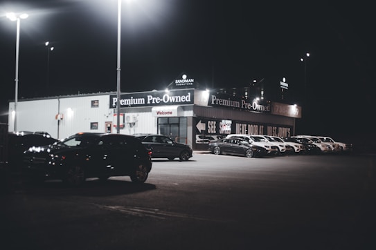 A dimly lit car dealership at night features a row of premium pre-owned cars parked in front of a building with bright lights. The building is adorned with various signs and has a 'Welcome' message visible. Multiple street lamps illuminate the parking lot.