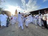 A group of people dressed in white are gathered in an outdoor, dusty area, engaged in a lively dance. Others are watching and clapping, forming a semi-circle around the dancers. In the background, a tall, decorated temple structure can be seen against a bright sky with scattered clouds.