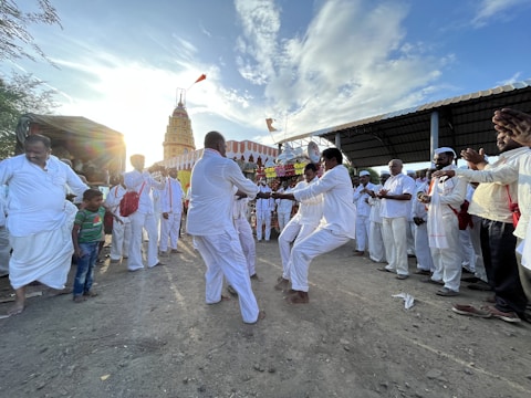 A group of people dressed in white are gathered in an outdoor, dusty area, engaged in a lively dance. Others are watching and clapping, forming a semi-circle around the dancers. In the background, a tall, decorated temple structure can be seen against a bright sky with scattered clouds.