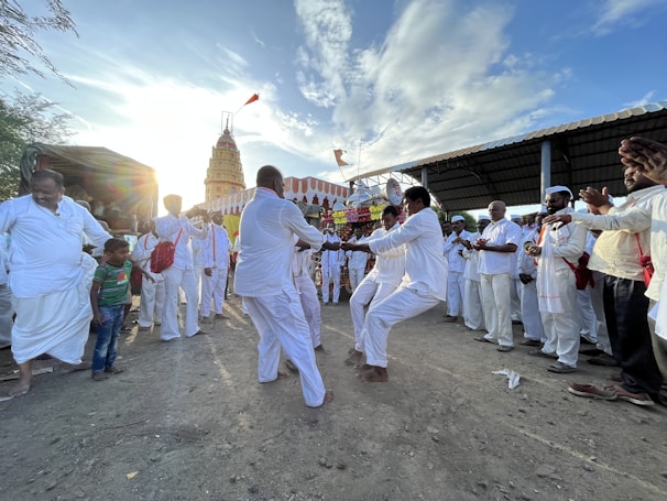 A group of people dressed in white are gathered in an outdoor, dusty area, engaged in a lively dance. Others are watching and clapping, forming a semi-circle around the dancers. In the background, a tall, decorated temple structure can be seen against a bright sky with scattered clouds.
