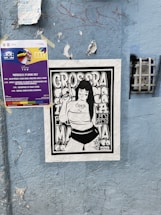 A blue wall features multiple posters. One is a colorful event poster with text about a performance scheduled for June 29, 2022. Below it, a black and white illustration of a person with long hair holding an object, surrounded by bold, graphic letters. The wall is textured and has some graffiti and peeling sections.
